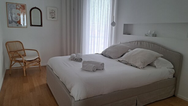 Ein ordentlich bezogenes Bett mit Kissen und aufgerollten Handtüchern, ein Korbstuhl und ein Bild an der Wand.