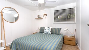 A bedroom with a bed, bedside tables, a mirror, a window with blinds, and a wall-mounted shelf with decorative items.