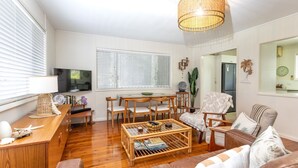 A living room with a TV, wooden furniture, a wicker coffee table, and a large window with blinds.
