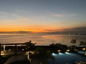 Udsigt over poolen med solnedgang over vandet, en by i det fjerne og en overdækket terrasse.