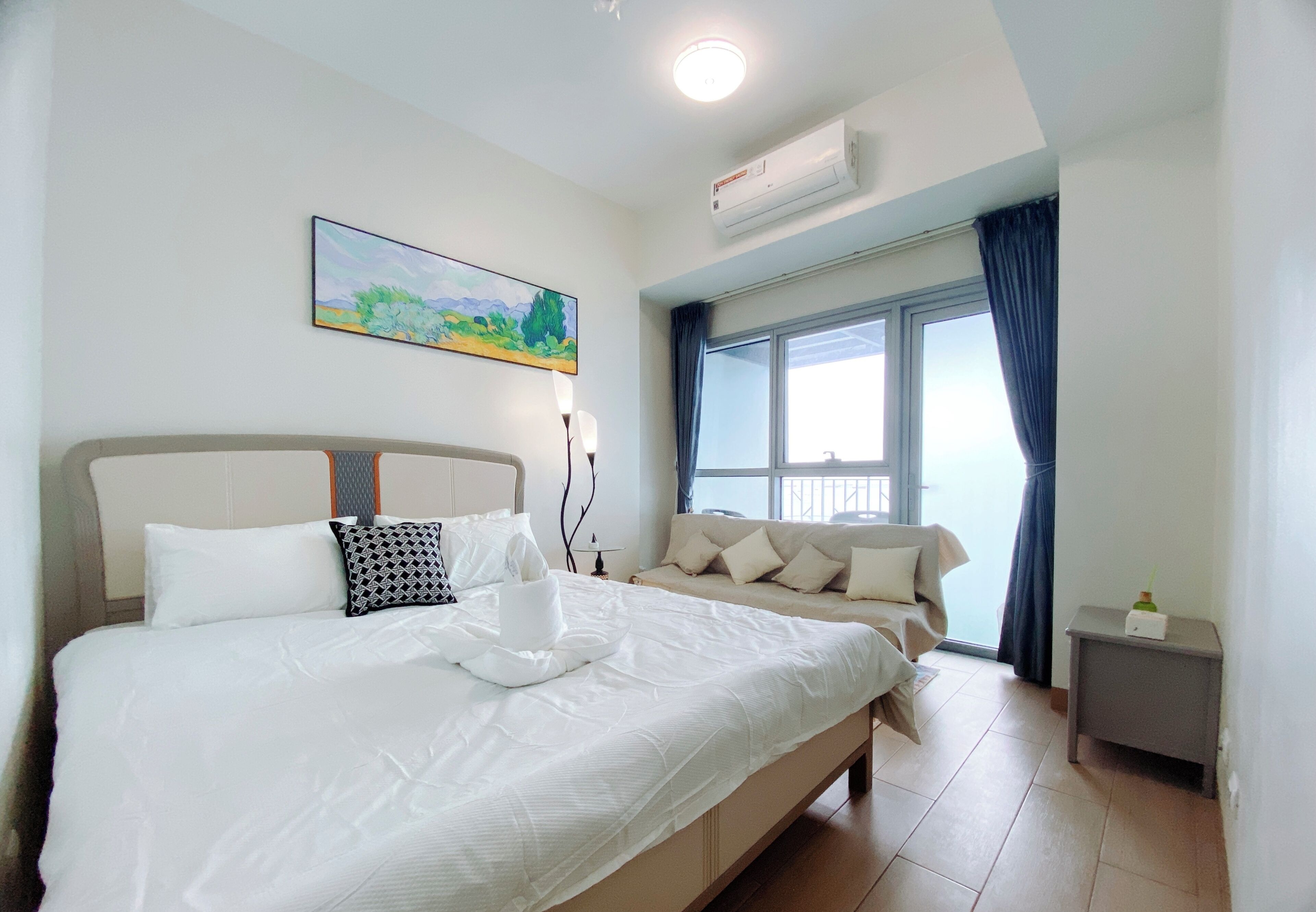 A hotel room with a large bed, a bedside table, a window with curtains, a painting on the wall, and an air conditioning unit.