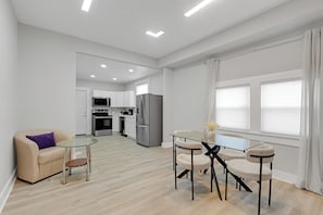 Eine moderne Küche mit Edelstahlgeräten, eine Essecke mit Glastisch und Stühlen sowie ein Wohnbereich mit einer Couch und einem kleinen Beistelltisch.