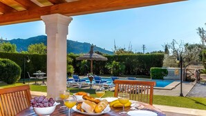 Una mesa de madera con un bol de uvas, un plato de pan y vasos de jugo de naranja, con vista a una piscina y montañas.