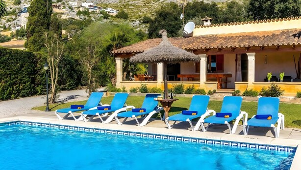 Una piscina con sillas reclinables azules, un quiosco con techo de paja y una casa con techo de tejas al fondo.