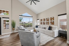 A spacious living room with a large window, a gray sofa, a coffee table, and a ceiling fan. - Tranquil 4-Bedroom House in charming Cave Creek-Fresh Remodel (Cave Creek)