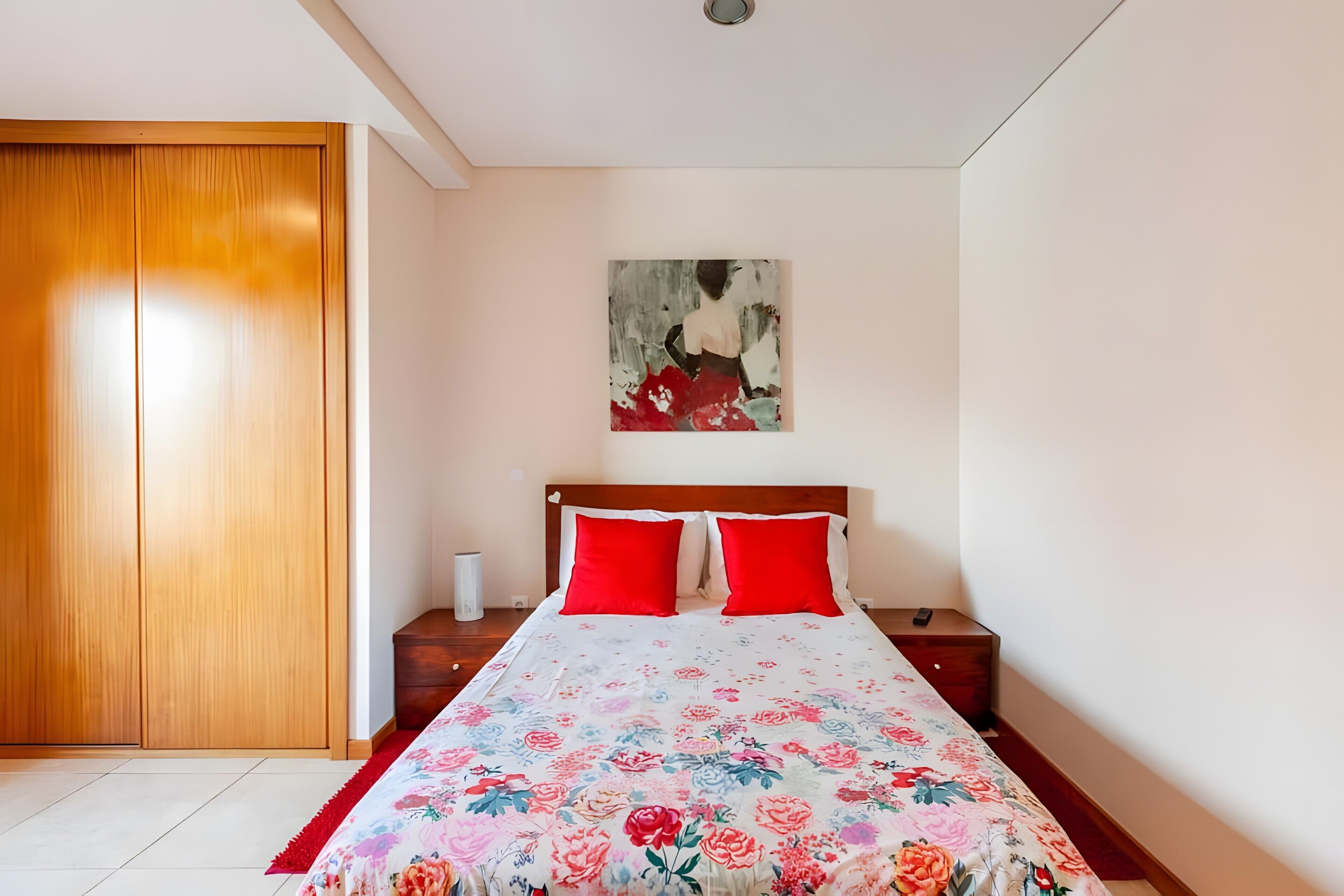 Un dormitorio con un armario de madera, una cama con colcha floral, dos almohadas rojas, una mesita de noche con lámpara y un cuadro en la pared.