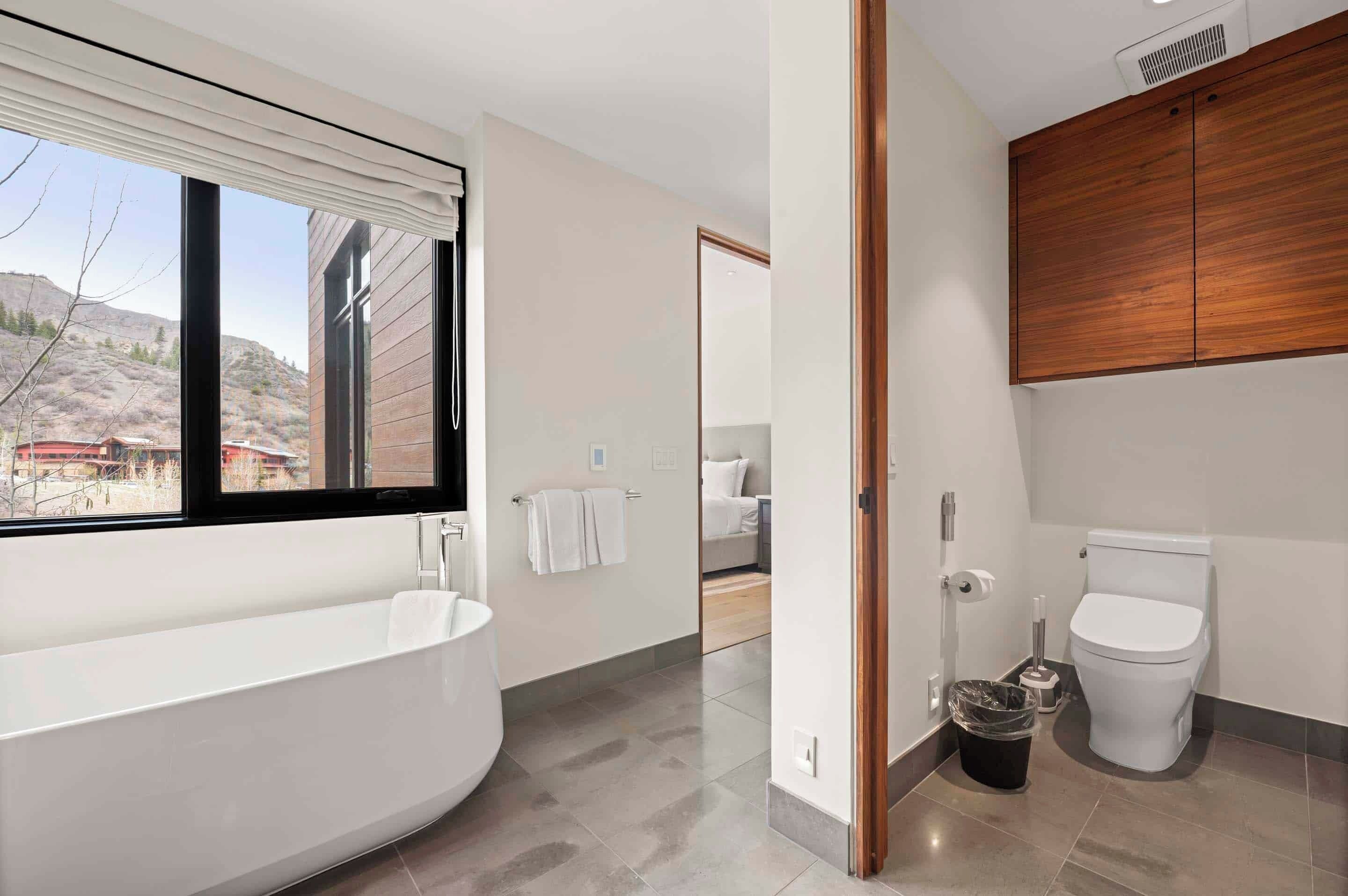 A modern bathroom with a bathtub, toilet, and a large window.