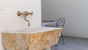 Un lavabo de piedra con grifo de latón, una silla y una mesa en una habitación con paredes blancas.