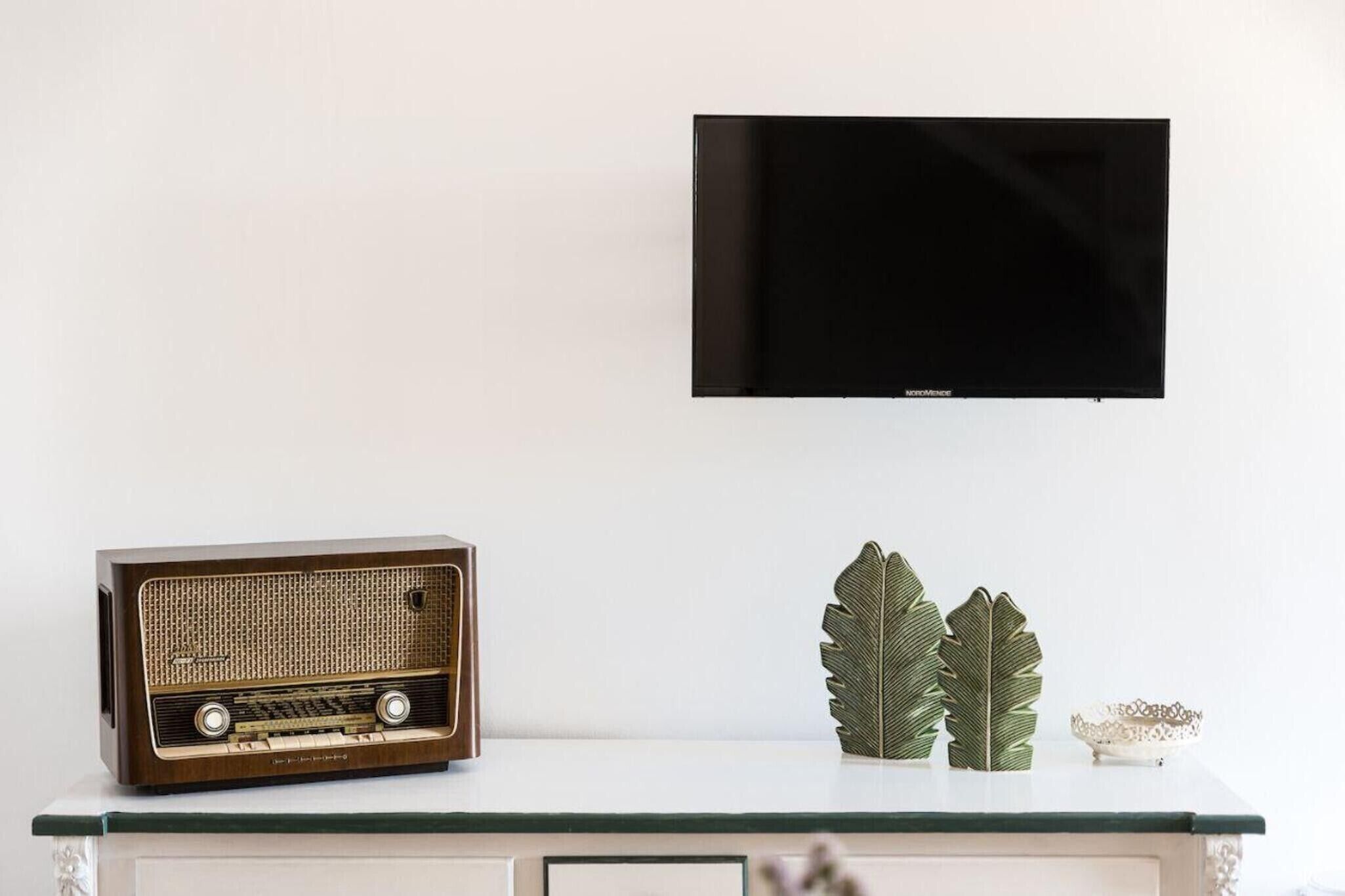 Un televisor de pantalla plana montado en la pared, una radio antigua sobre una mesa de vidrio, y hojas decorativas con un tazón.