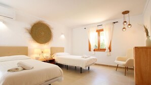 Quarto de hotel com duas camas, uma cômoda de madeira, um ornamento circular na parede, uma janela com cortinas e um lustre.