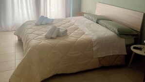 Una cama bien hecha con cubrecolcha acolchada, una mesita de noche con lámpara, y una ventana con cortinas traslúcidas.