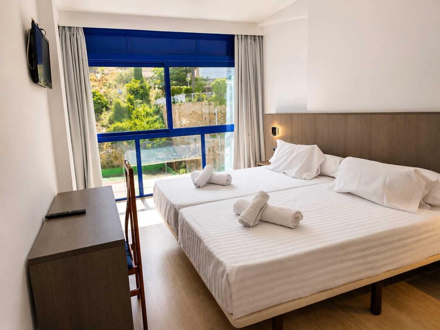 A hotel room with two beds, a desk, and a large window with a view of greenery.