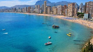Paisagem costeira com praia, barcos e edifícios altos.