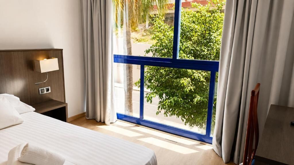 A hotel room with a bed, a nightstand, a lamp, a window with curtains, and a view of greenery outside.
