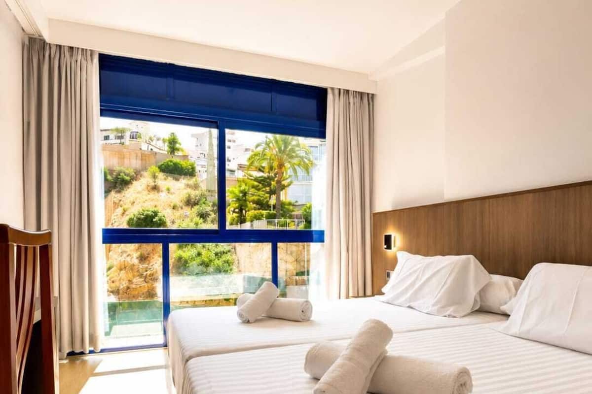 A hotel room with two beds, a wooden headboard, a view of a hillside, and a balcony.