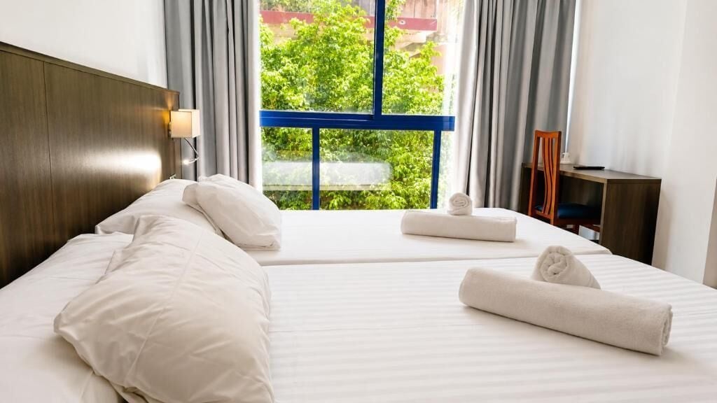 A hotel room with a bed, a nightstand, a lamp, a window with curtains, and a view of greenery outside.