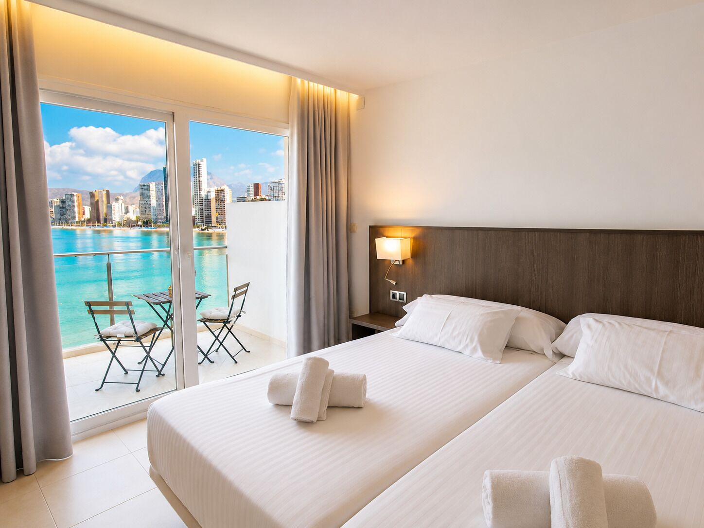 Una camera d'albergo con un letto, un comodino, una lampada e vista sulla città e sul mare dalla finestra.