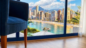 Quarto com grande janela com vista para a praia e paisagem urbana.