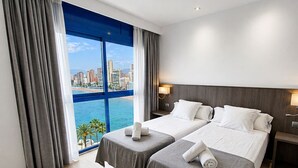 Quarto de hotel com duas camas, uma janela grande com vista para a cidade e uma mesa de cabeceira com abajur.