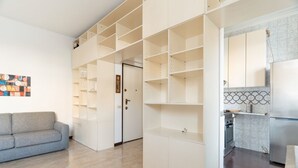 A modern living space with a grey sofa, a built-in shelving unit, a kitchen area with a stove and refrigerator, and a door leading to another room. - GuestHost - Cozy apartment of approximately 55 m2, able to accommodate up to 4 people, located on the first floor of a building with lift (NOT suitable for people in wheelchairs).The property is located in the Risorgimento district, in a residential area (Milan)