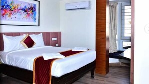 A hotel room with a bed, a ceiling fan, an air conditioning unit, a large window with curtains, and a colorful abstract painting on the wall. - Narayani residency (Tiruvannamalai)