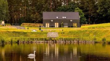 Ein Haus mit dunklem Dach und weißen Fenstern, umgeben von einer Wiese und einem Gewässer mit einem Schwan.