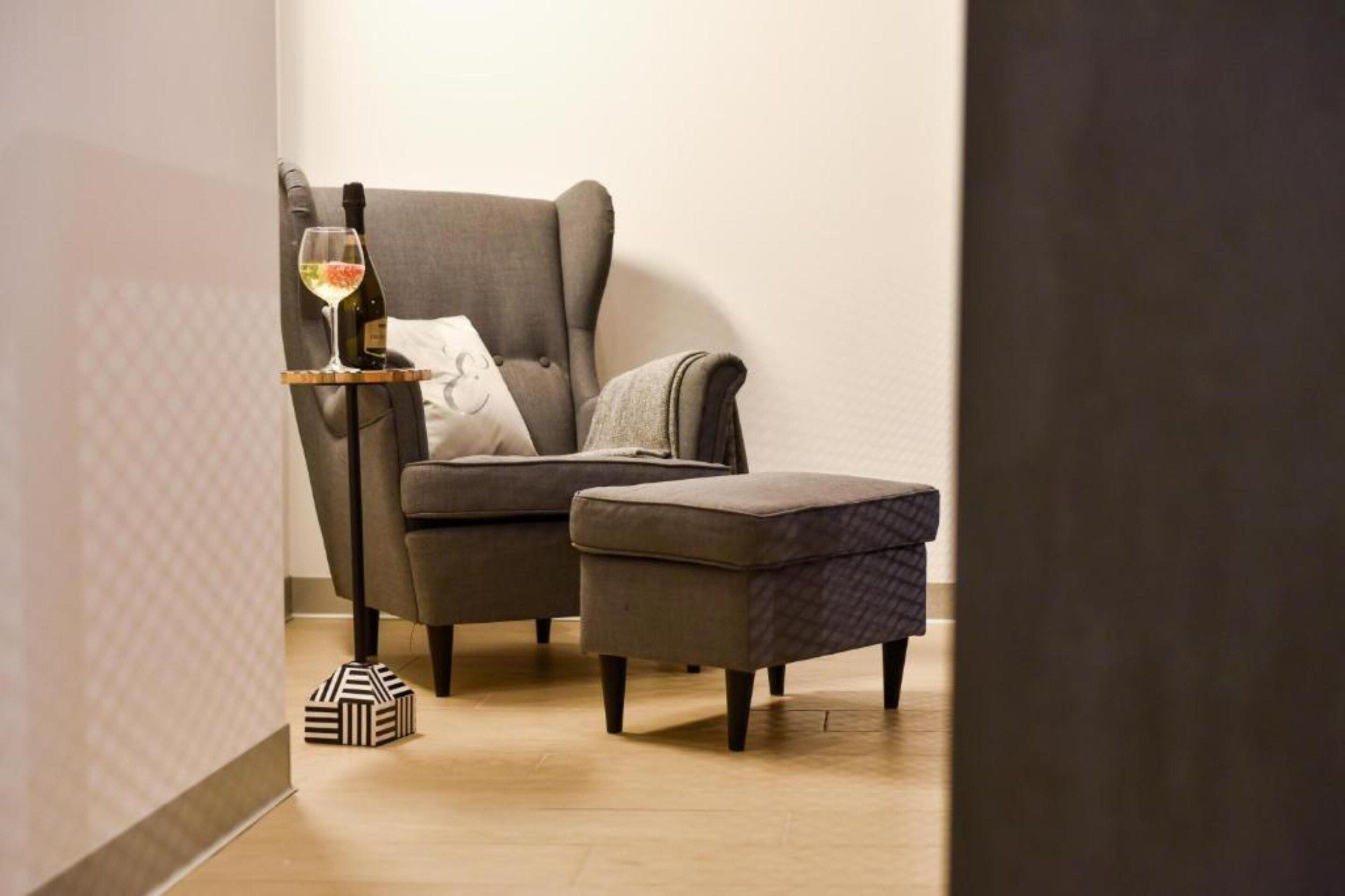 A room with a grey armchair, a small ottoman, a side table with a wine glass and bottle, and a patterned bag on the floor.