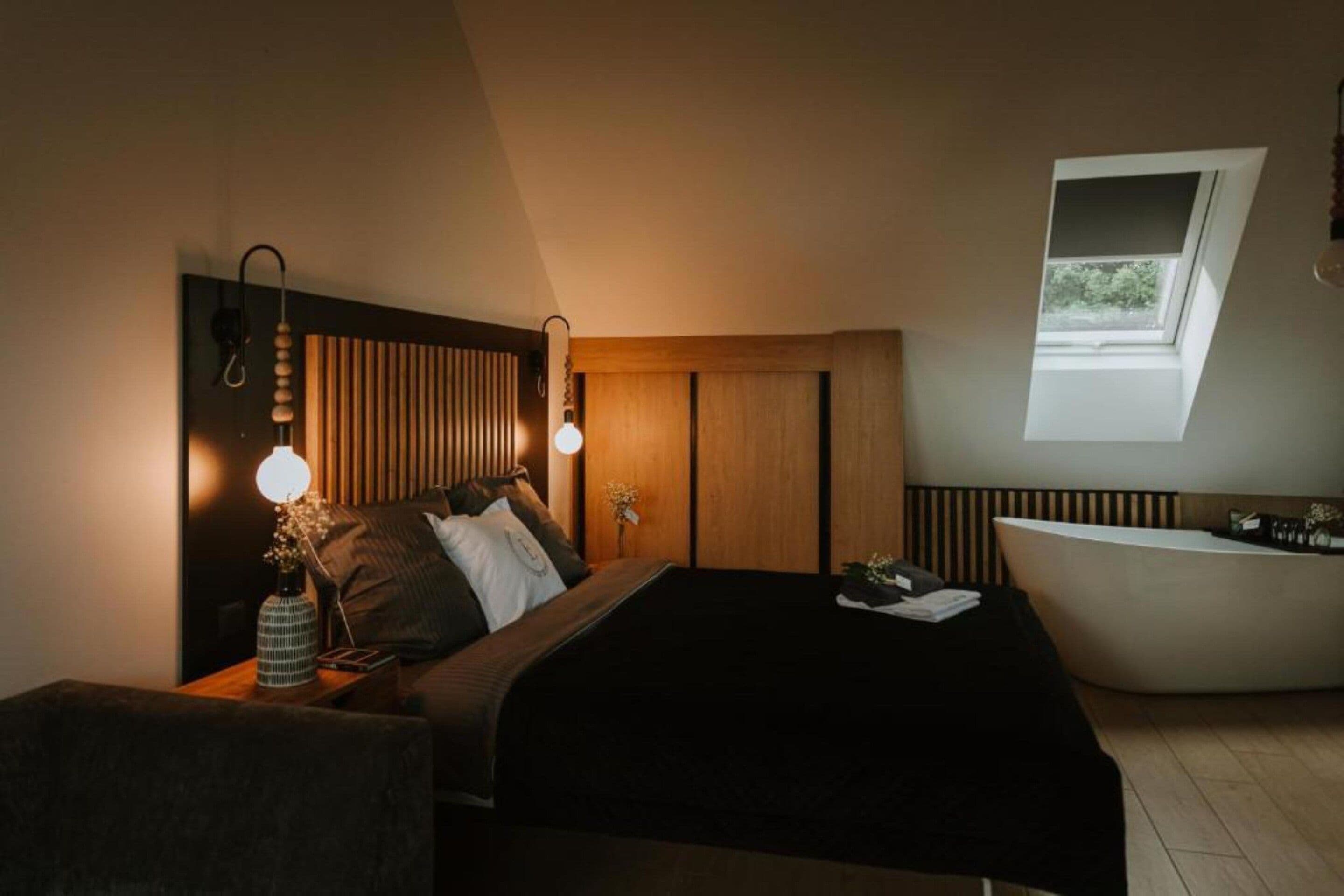 A bedroom with a bed, bedside lamps, a nightstand, a window, and a bathtub.