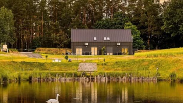 A building with a dark roof and multiple windows, surrounded by a grassy area and a body of water with a swan. - Holiday Home, Bedroom With Bathtub, Jacuzzi (Koscierzyna)
