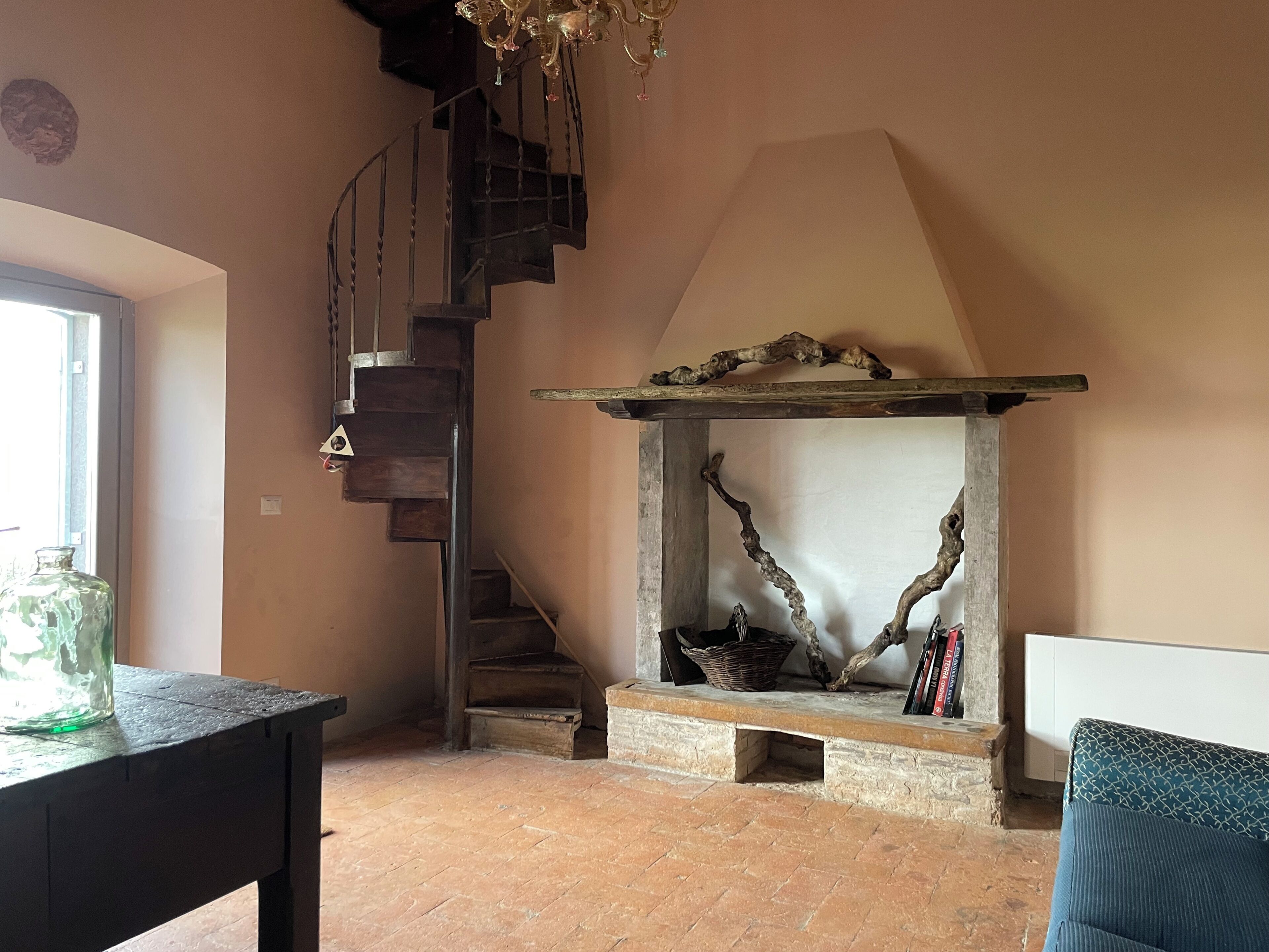 A room with a spiral staircase, a fireplace with a rustic design, and a window providing natural light.