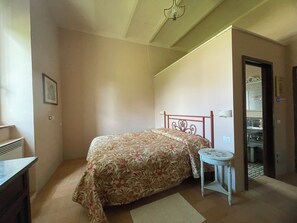 A bedroom with a bed, a small table, a chair, a dresser, and a bathroom visible through an open door. - Olive Tree Suite (Sugano)
