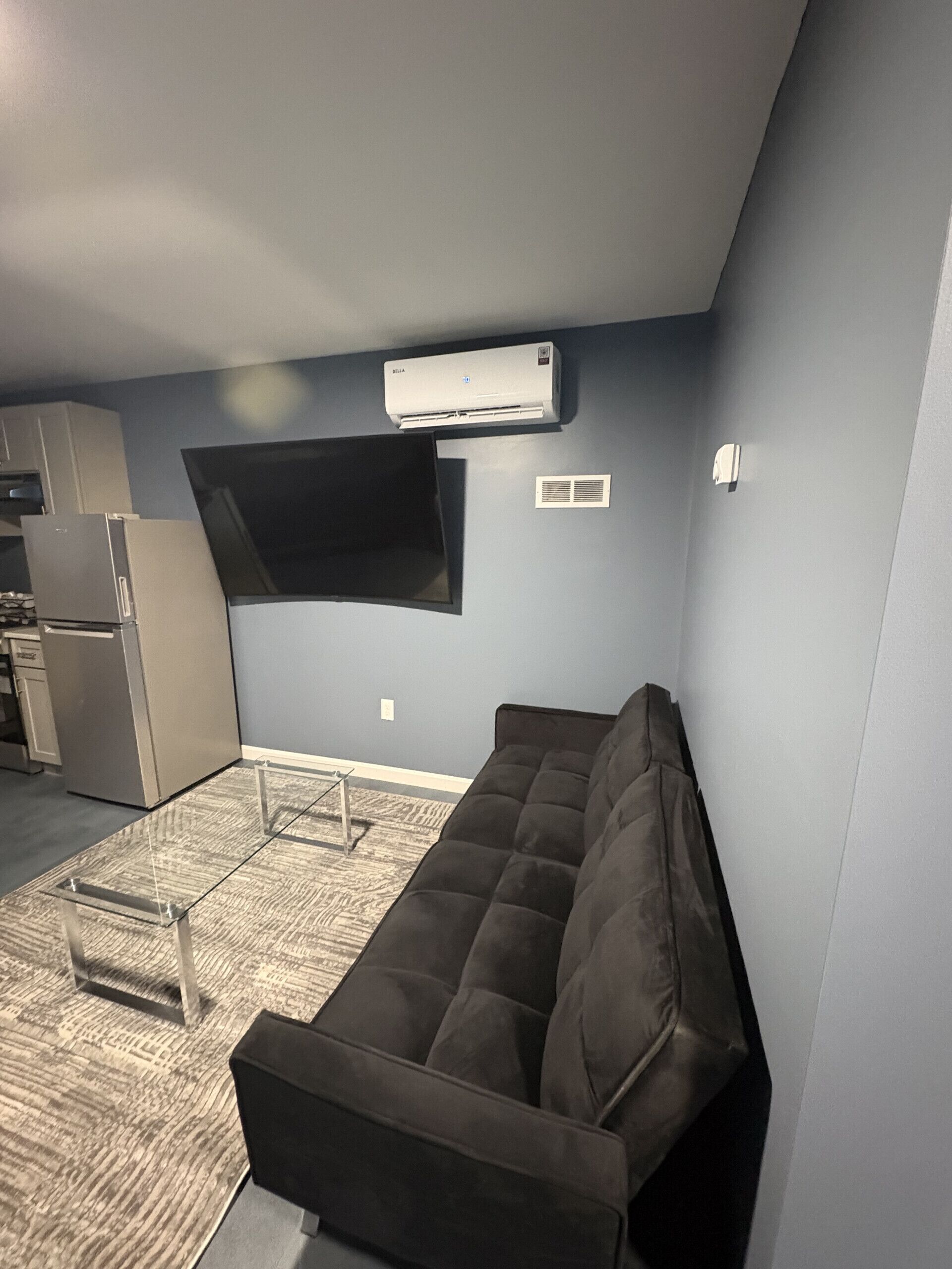 A compact living space with a sofa, a flat-screen TV mounted on the wall, a built-in air conditioner, and a kitchenette area.