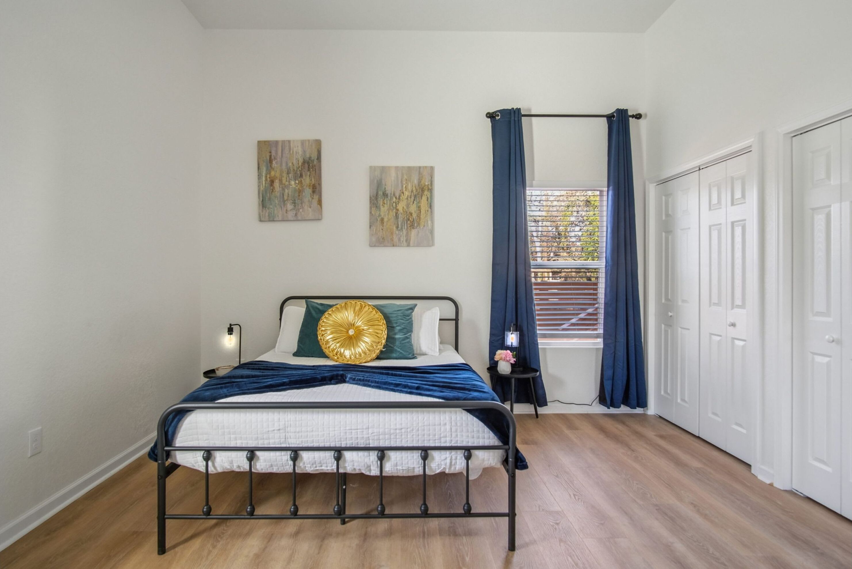 Una camera da letto con un letto, due opere d'arte a parete, una finestra con tende, un comodino e una porta.