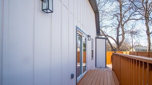 Una terraza de madera que conduce a un edificio blanco con puerta corredera de vidrio y una estructura pequeña adosada.