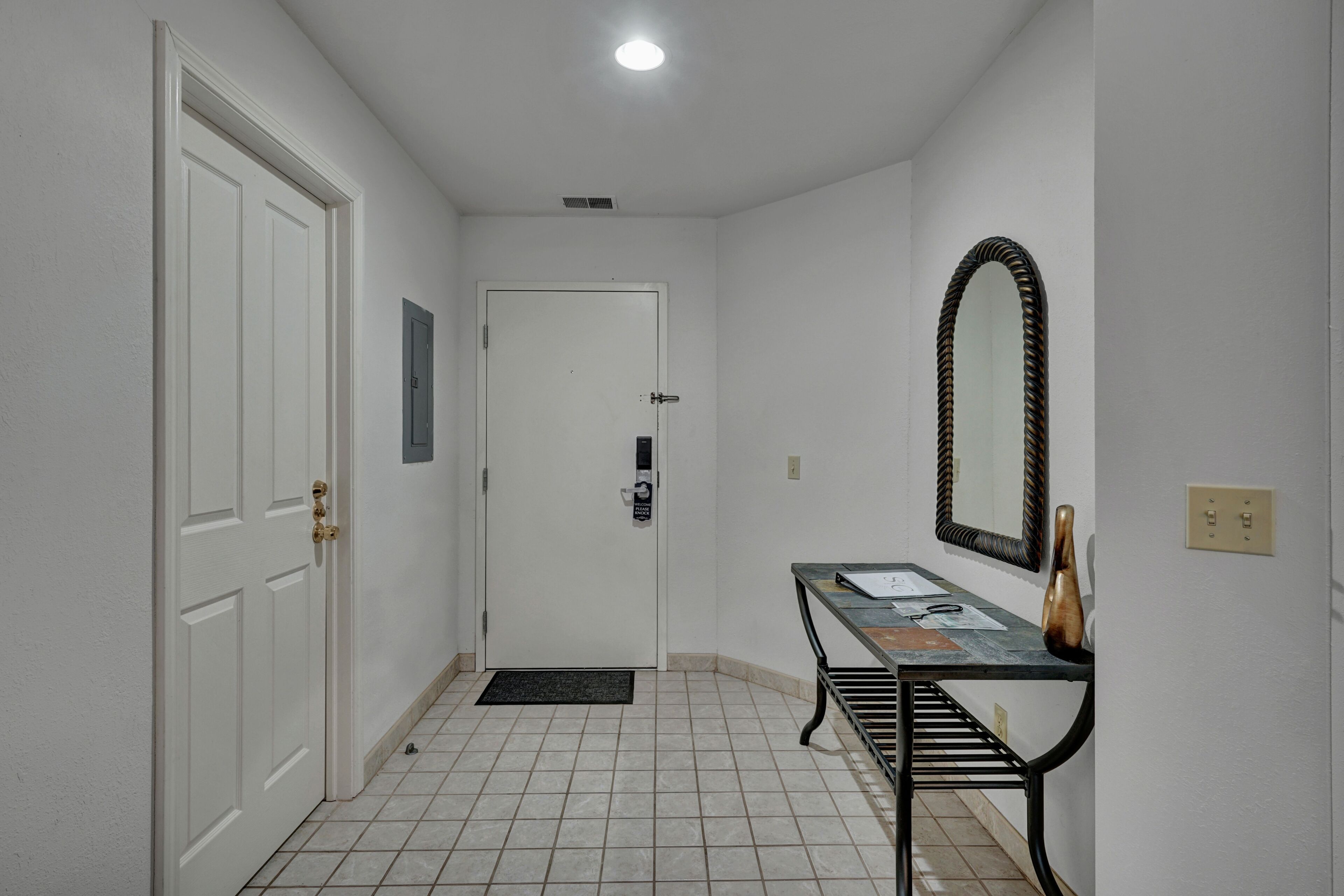 A hallway with a white door, a small table with a mirror, and a tiled floor.