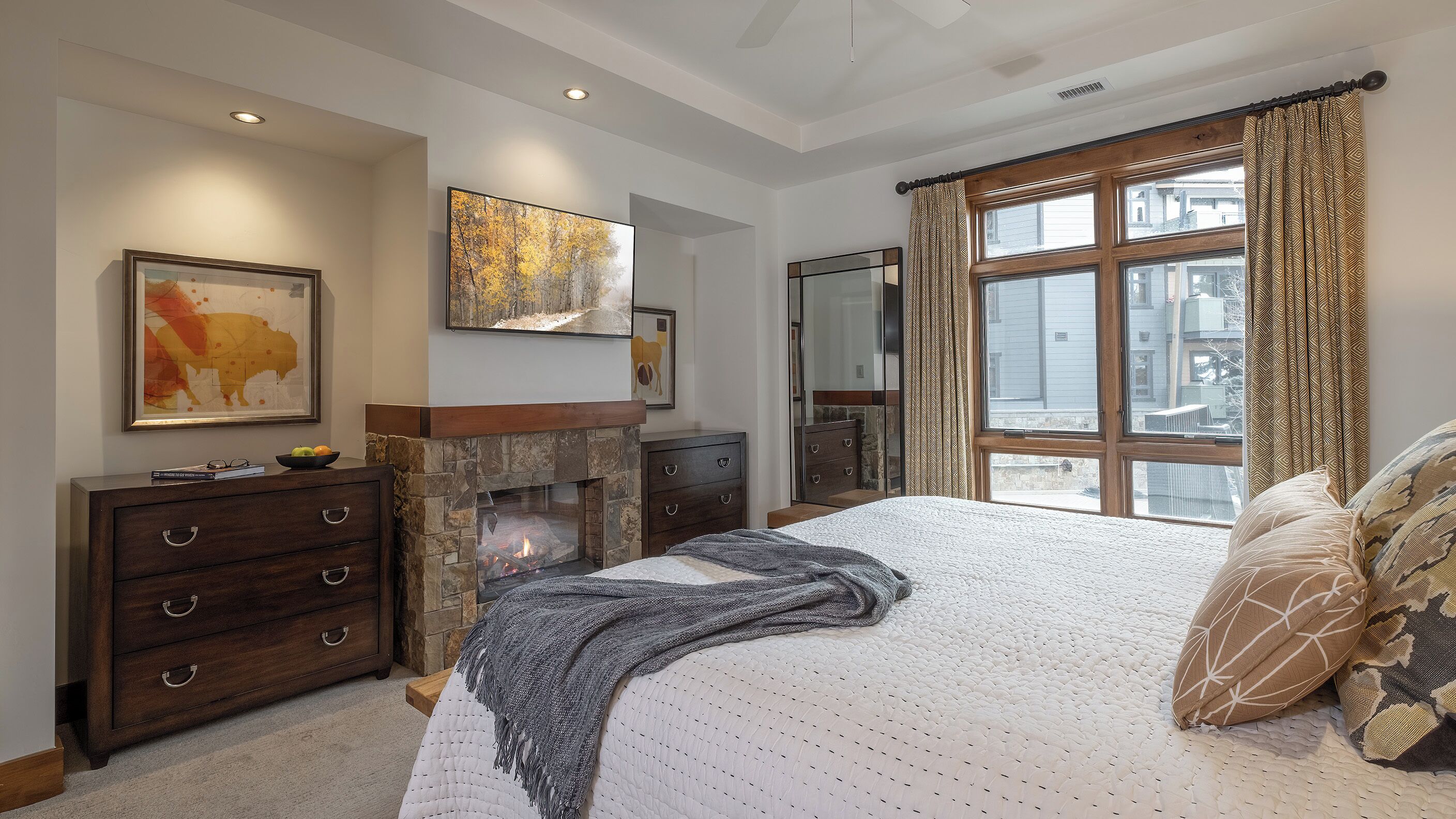 Une chambre avec une cheminée en pierre, un grand lit agrémenté d’oreillers à motifs et une vue sur les bâtiments, visible à travers une fenêtre ornée de rideaux à motifs.