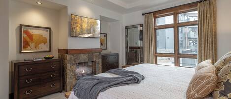 Une chambre avec une cheminée en pierre, un grand lit agrémenté d’oreillers à motifs et une vue sur les bâtiments, visible à travers une fenêtre ornée de rideaux à motifs.