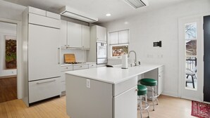 A modern kitchen with white cabinetry, a central island with stools, a stove, and a window with blinds. - MINNeSTAY* Como Classic (St. Paul)