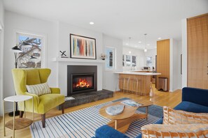 Ein modernes Wohnzimmer mit Kamin, einem gelben Sessel, einem blauen Sofa und einer Küchenzeile im Hintergrund.