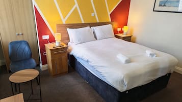 A hotel room with a bed, a desk, a chair, a nightstand with a lamp, and a wall with a geometric design.
