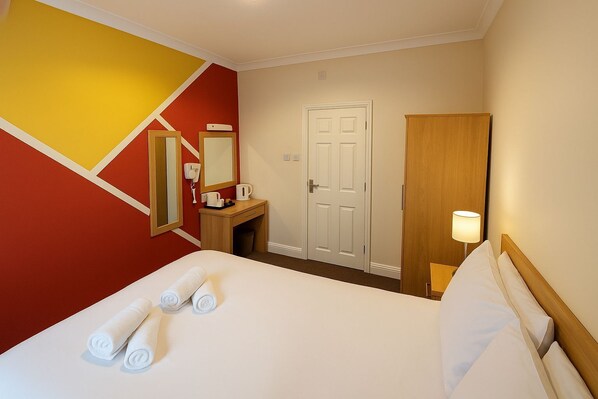 A hotel room with a bed, a desk with a telephone and a coffee maker, a mirror, a door, and a nightstand with a lamp.