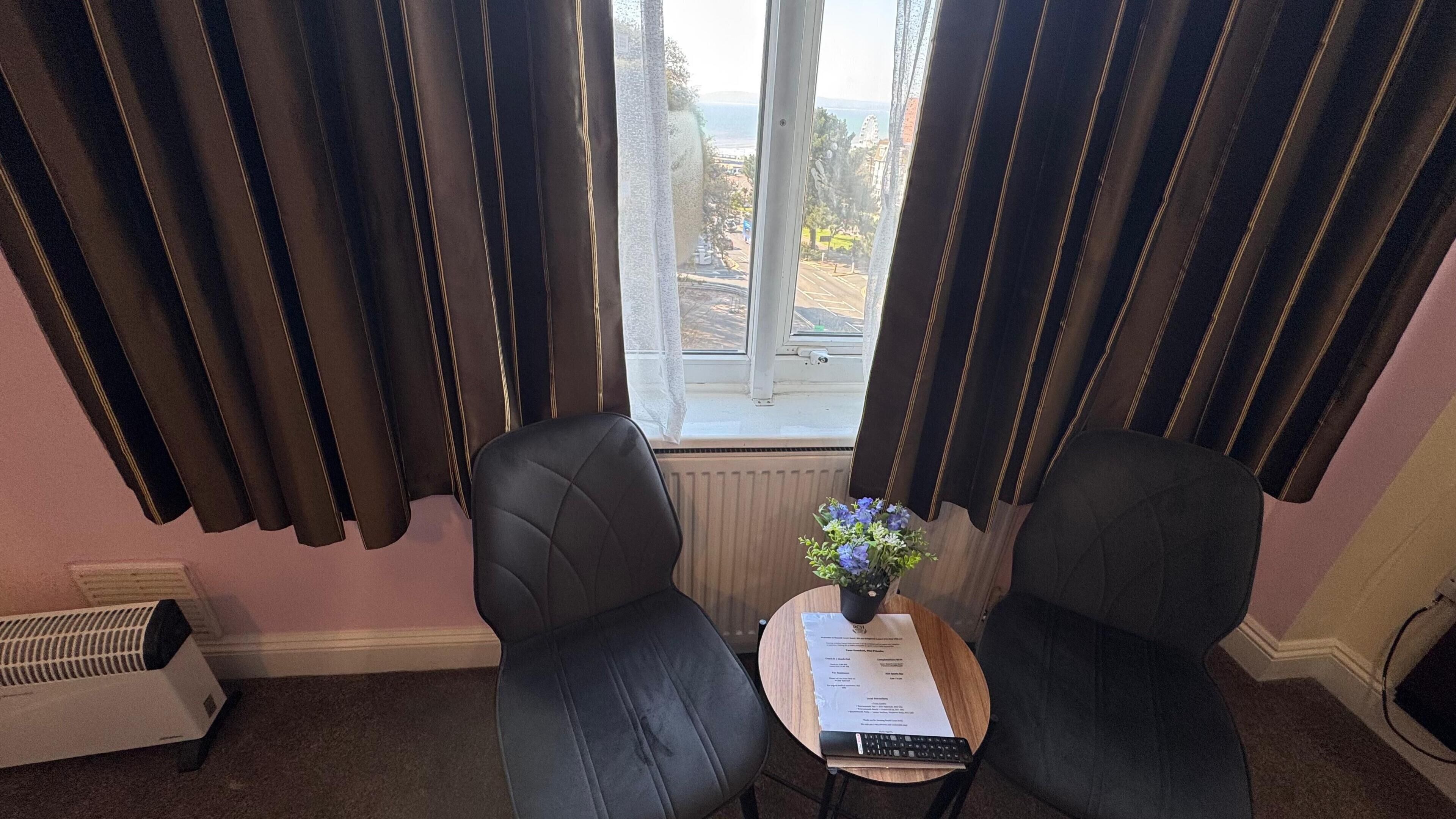 A room with two black chairs, a small table with a vase of flowers, a radiator, and a window with curtains.