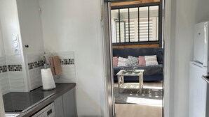 Una cocina compacta con lavavajillas, un cesto de basura y vistas hacia una sala de estar con sofá y ventana.