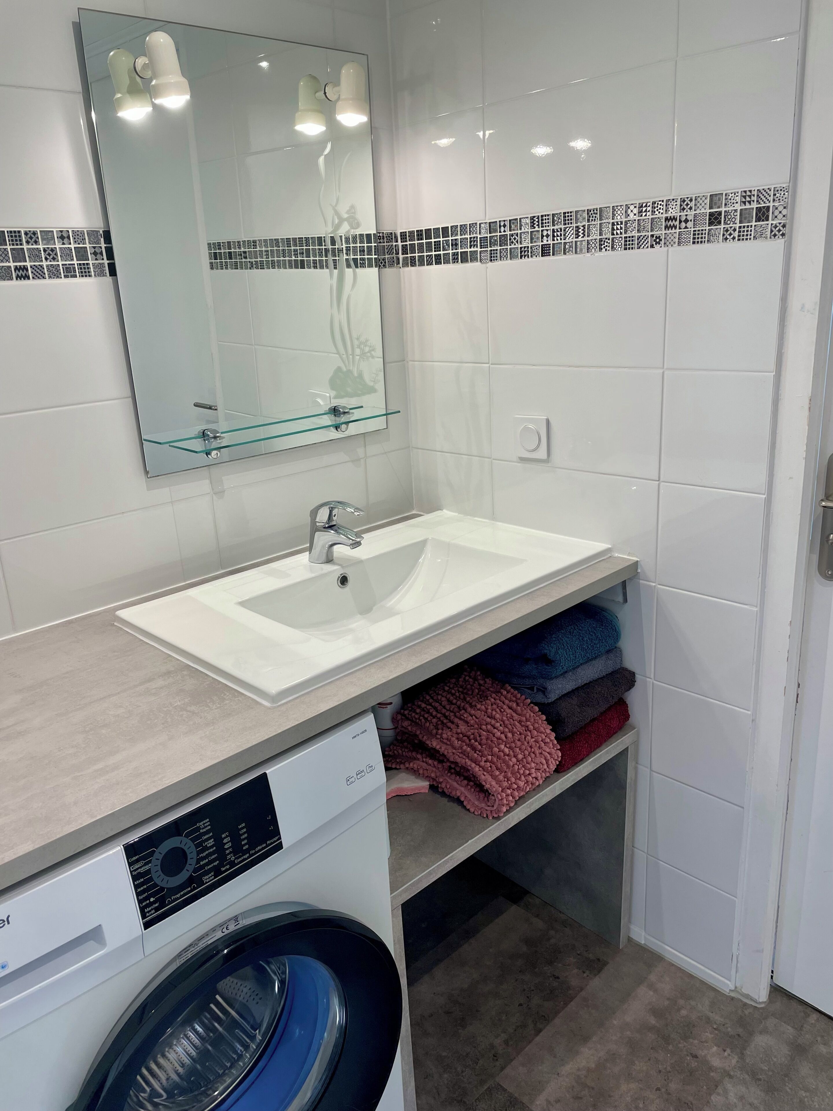 A bathroom with a white sink, a glass shelf, a washing machine, and a mirror with a decorative tile border.