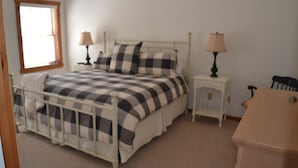 Une chambre à coucher avec un lit en métal, une couvre-lit à carreaux, deux tables de chevet avec des lampes, une commode en bois et une fenêtre avec des stores.