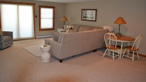 Un salon avec un canapé beige, une petite table ronde et deux chaises, des toilettes blanches et une fenêtre avec des stores.