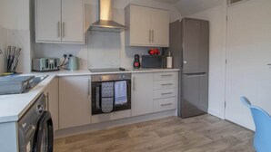 A compact kitchen with white cabinets, a stainless steel range hood, a built-in oven, a refrigerator, and a dishwasher. - Ash House 3 Bedroom in Cumbernauld (Glasgow)