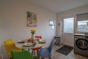 Een eetgedeelte met een ronde tafel, kleurrijke stoelen, een vaas met bloemen en een wasmachine in de hoek.