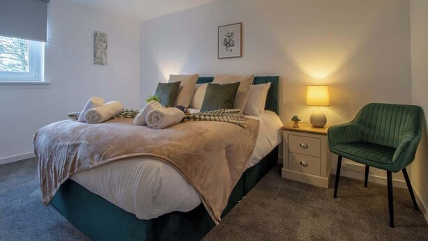 A bedroom with a bed, bedside table, lamp, chair, and artwork on the wall. - Ash House 3 Bedroom in Cumbernauld (Glasgow)