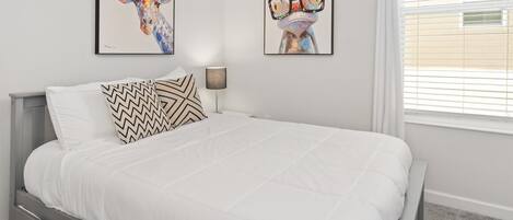 Een slaapkamer met een bed, twee kunstwerken aan de muur, een bedlampje en een raam met zonwering.
